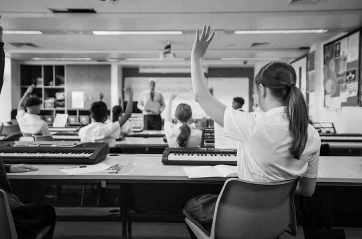 A wide shot of a classroom with a one student raise their hand for the teacher at the front of the class